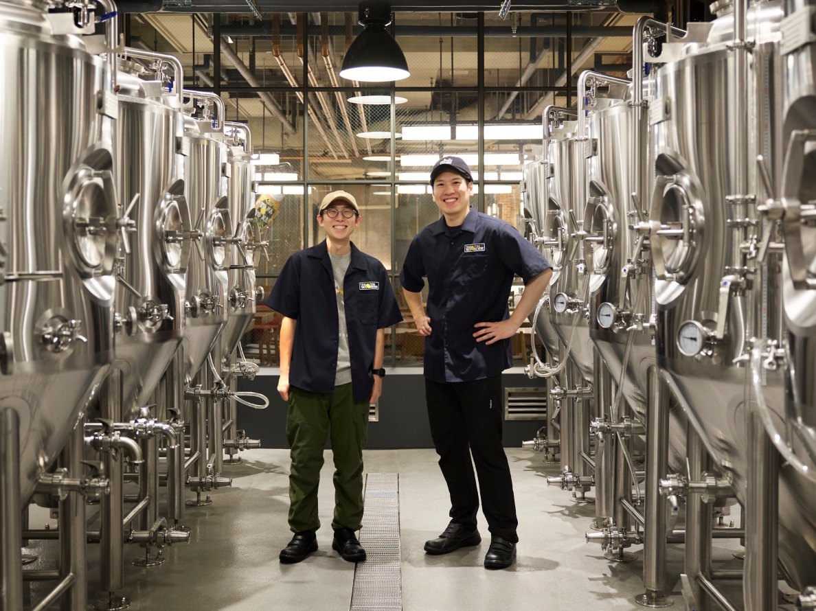 YONA YONA TOKYO BREWERY、醸造タンクが両脇に並ぶ通路、笑顔を見せる二人の醸造スタッフ