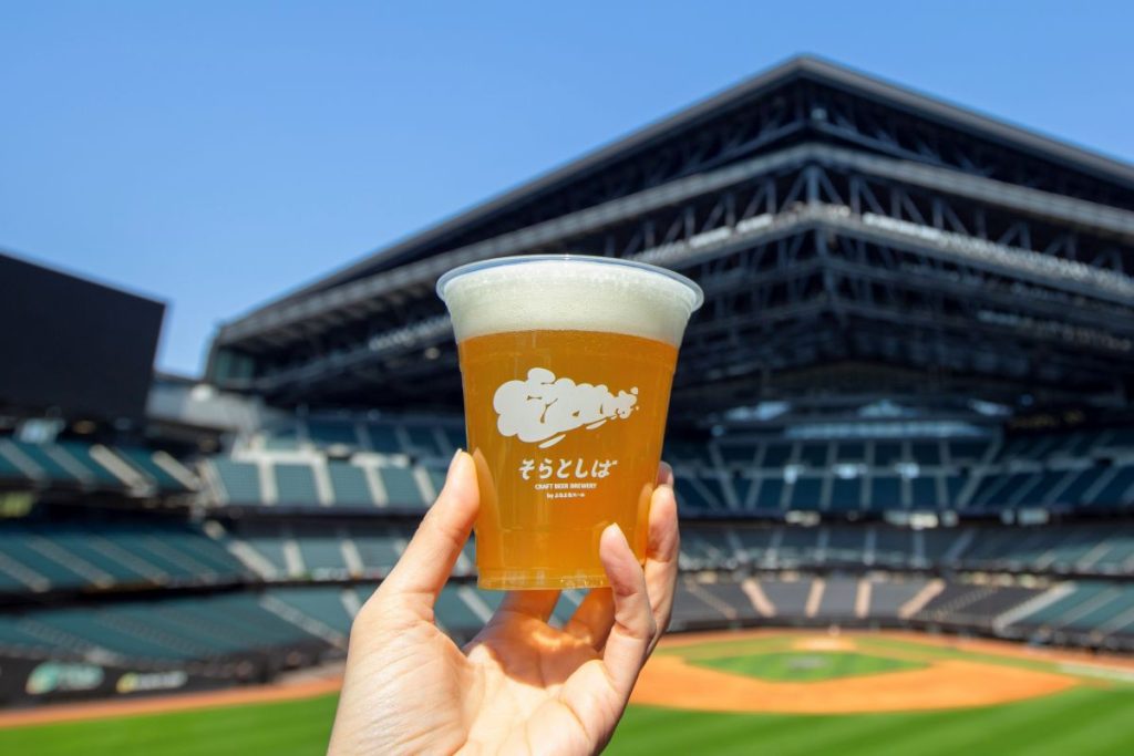 エスコンフィールドHOKKAIDOをバックにクラフトビール「そらとしば Play Ball! Ale」を掲げている様子